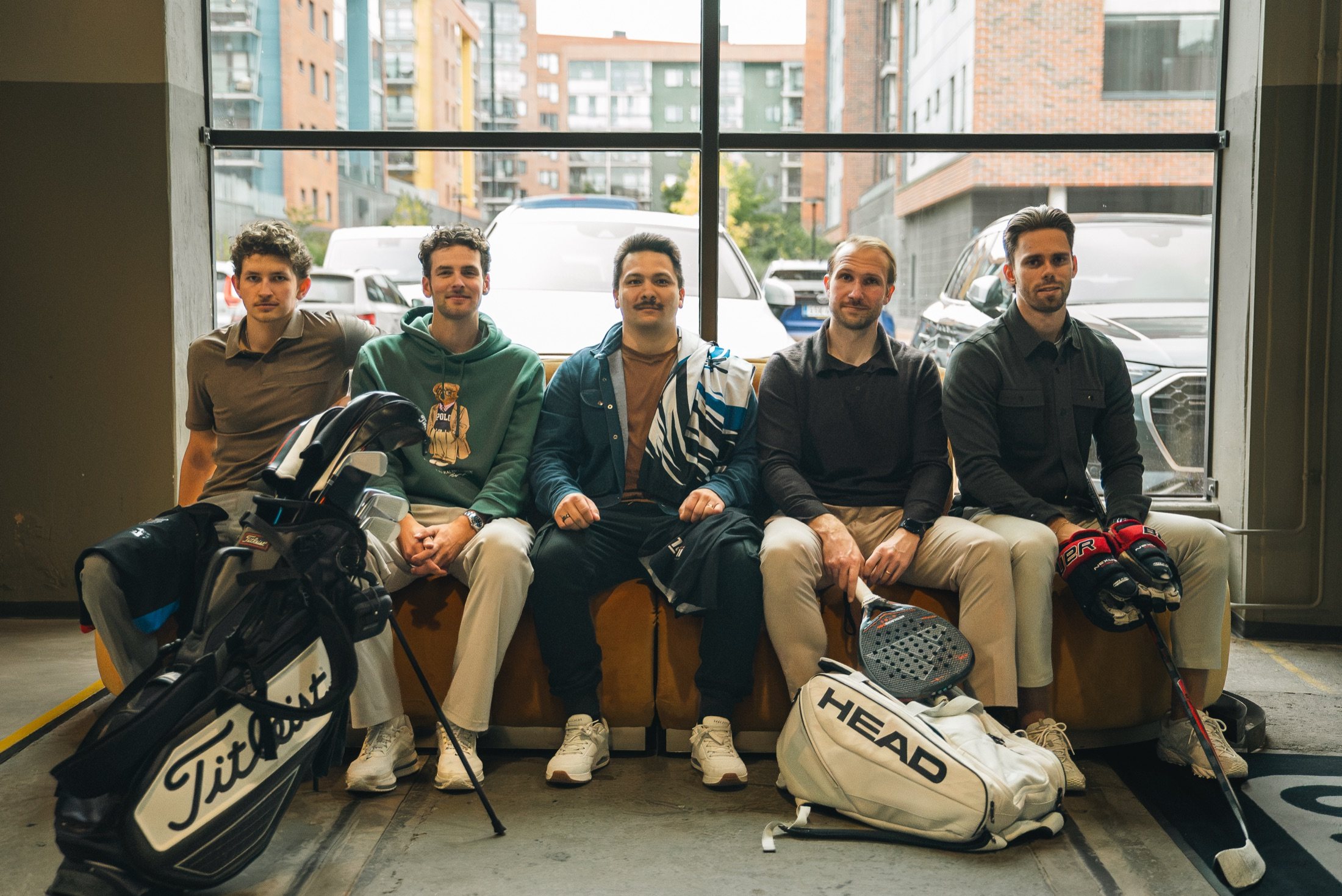 The Innovila team on a sofa with their sports gear: a golf bag, a padel racket and bag, and ice hockey gloves and a stick
